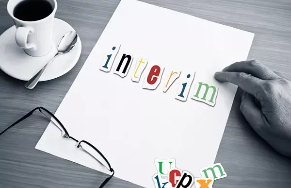 Hand holding paper with 'interim' spelled in colorful letters, next to glasses and a coffee cup.