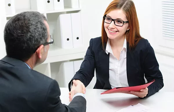 Two people shaking hands at a desk during a meeting.